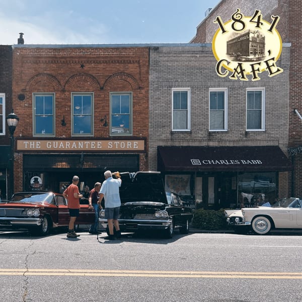 Classic cars parked on a street in front of 1841 Cafe and other brick buildings.