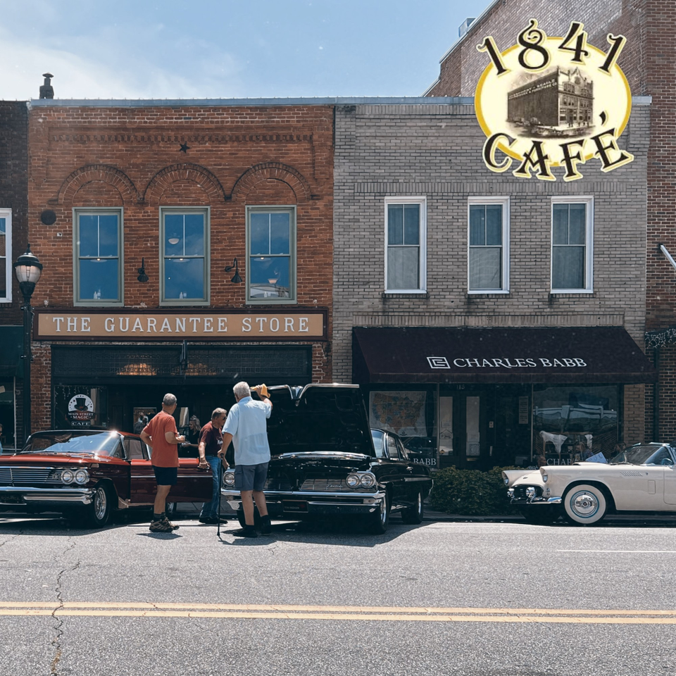 Classic cars parked on a street in front of 1841 Cafe and other brick buildings.