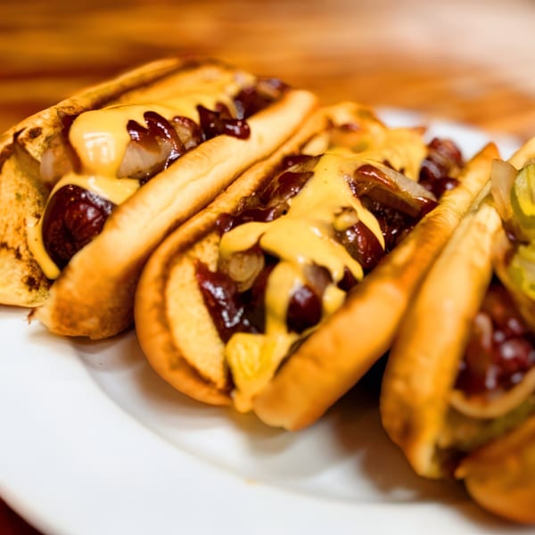 Three hot dogs in buns, topped with melted cheese, caramelized onions, and barbecue sauce.