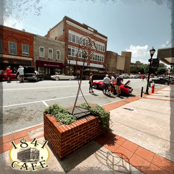 A street view of 1841 Cafe with classic cars and people on a sunny day.
