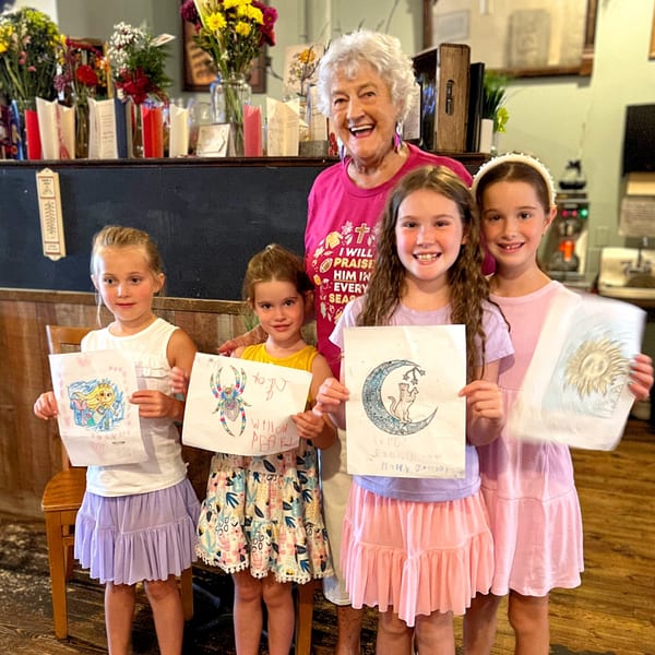 A smiling older woman and four young girls proudly display their colorful drawings in a restaurant.