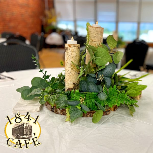 A rustic centerpiece with birch logs, candles, and various green foliage on a white tablecloth.