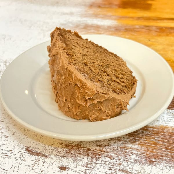 A slice of chocolate cake with chocolate frosting on a white plate, on a distressed wooden table.