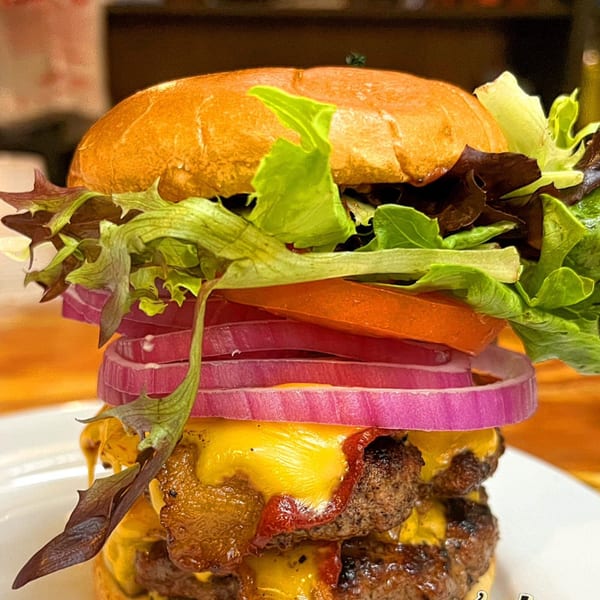 A towering double cheeseburger with bacon, red onion, tomato, and lettuce on a brioche bun.