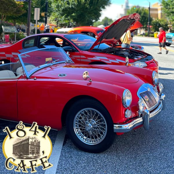 Two red classic cars, one convertible, parked on a street with people in the background.