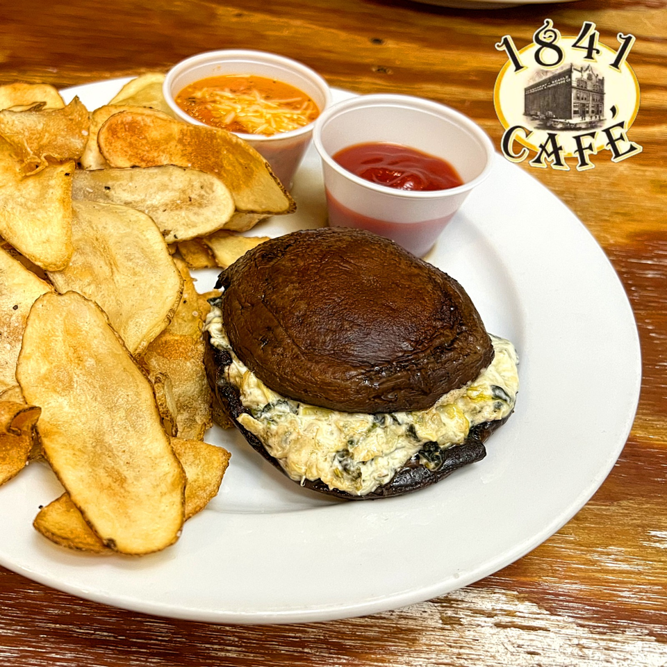1841 Cafe burger with creamy filling, homemade potato chips, and cups of tomato dipping sauces on a white plate.