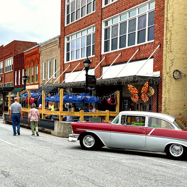 A red and silver vintage car parked on a street with people walking past 1841 Cafe's outdoor seating.