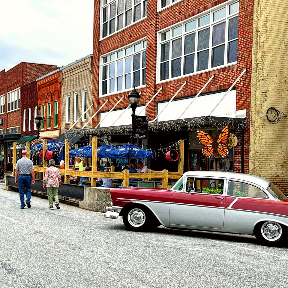 A red and silver vintage car parked on a street with people walking past 1841 Cafe's outdoor seating.