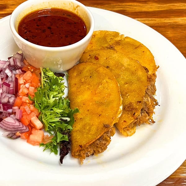 Three golden birria tacos with a side of consommé, diced red onions, tomatoes, and lettuce.