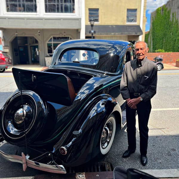 An older man stands proudly beside a shiny black vintage car with whitewall tires on a sunny street.
