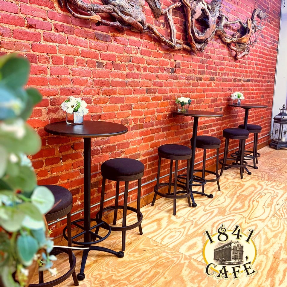 Tall tables with stools line a brick wall adorned with wooden art at 1841 Cafe.