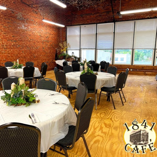 A spacious event room with round tables, white tablecloths, and black chairs, featuring brick walls and large windows.