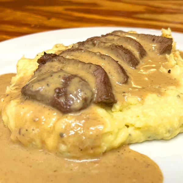 Sliced meat with gravy on a bed of mashed potatoes, served on a white plate.