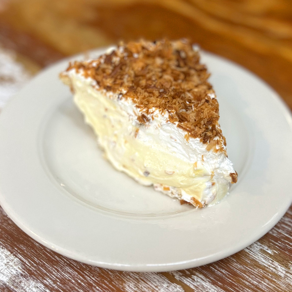 Slice of creamy layered cake topped with toasted coconut flakes on a white plate.
