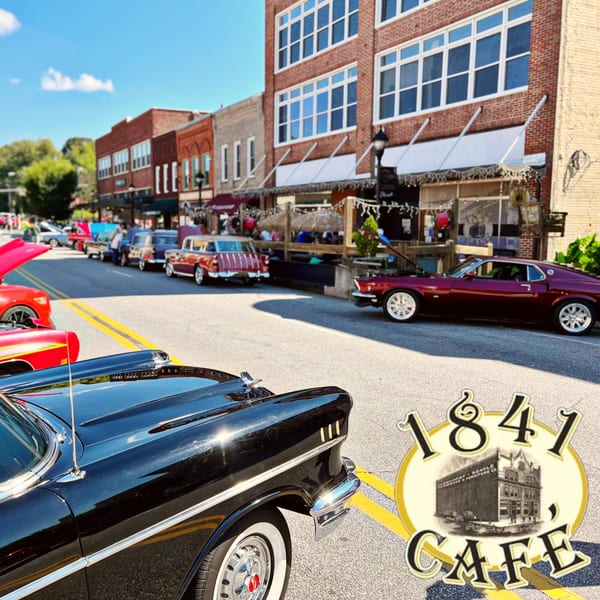 Classic cars parked along a street in front of 1841 Cafe and other brick buildings.