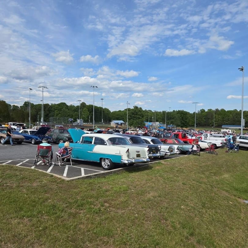 Classic car show in a parking lot with many vintage vehicles and people enjoying the sunny day.