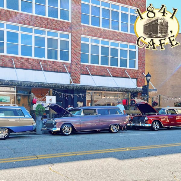 Classic cars parked on the street in front of 1841 Cafe, a brick building with awnings.