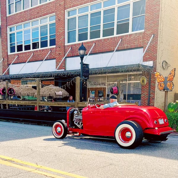 A red hot rod drives past 1841 Cafe's outdoor patio with tiki umbrellas and string lights.