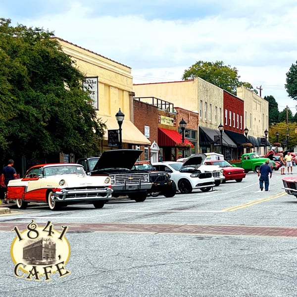 A street lined with classic cars and storefronts, with people walking around during a car show.