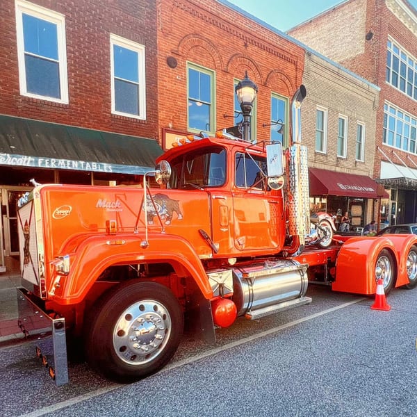 A bright orange Mack truck with chrome details parked on a street in front of brick buildings.