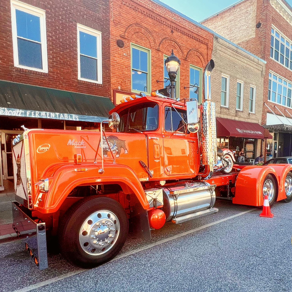 A bright orange Mack truck with chrome details parked on a street in front of brick buildings.
