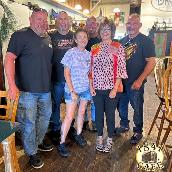 Six smiling people, three men and three women, stand together in a rustic cafe interior.