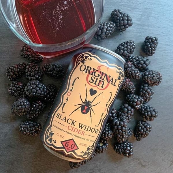 Original Sin Black Widow Cider can surrounded by fresh blackberries and a glass of red liquid.