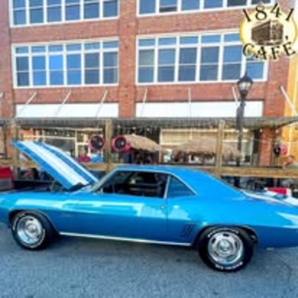 A blue classic car with its hood open parked outside the 1841 Cafe, with people on the patio.