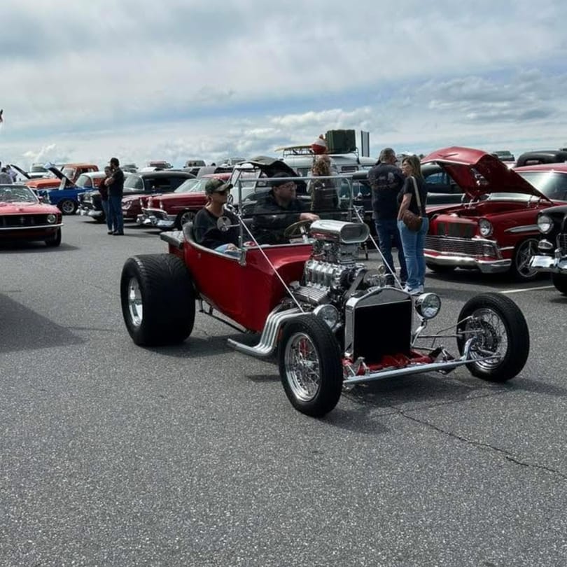Red hot rod with exposed engine at an outdoor car show with people and other classic cars.