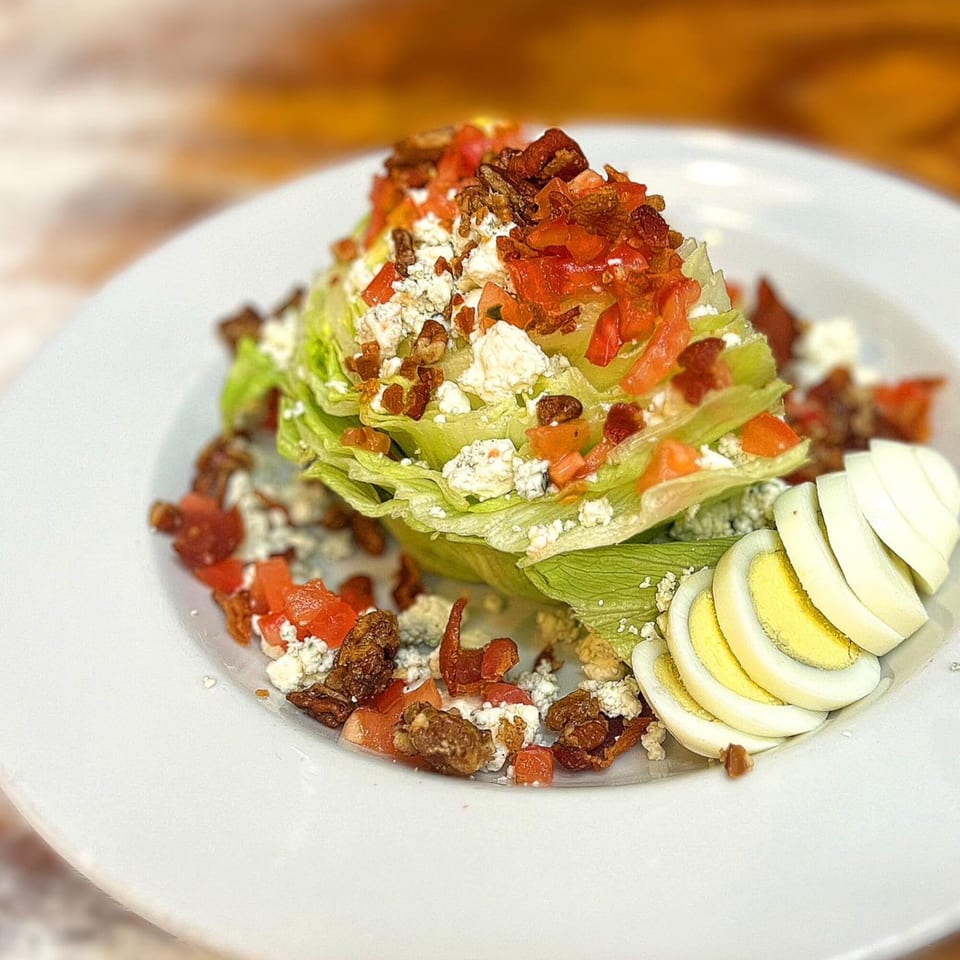 A wedge salad with blue cheese, bacon, tomatoes, and sliced hard-boiled eggs on a white plate.