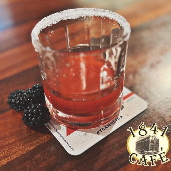 A red cocktail with a sugared rim and blackberries on a coaster with the 1841 Cafe logo.