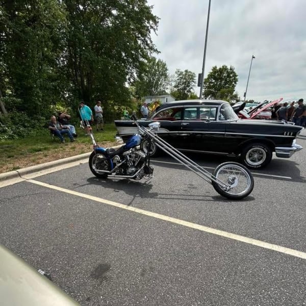 Custom blue chopper motorcycle with extended forks parked next to a black classic car at an outdoor event.
