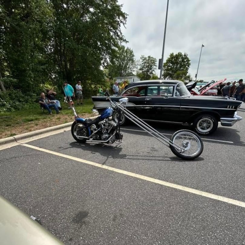 Custom blue chopper motorcycle with extended forks parked next to a black classic car at an outdoor event.