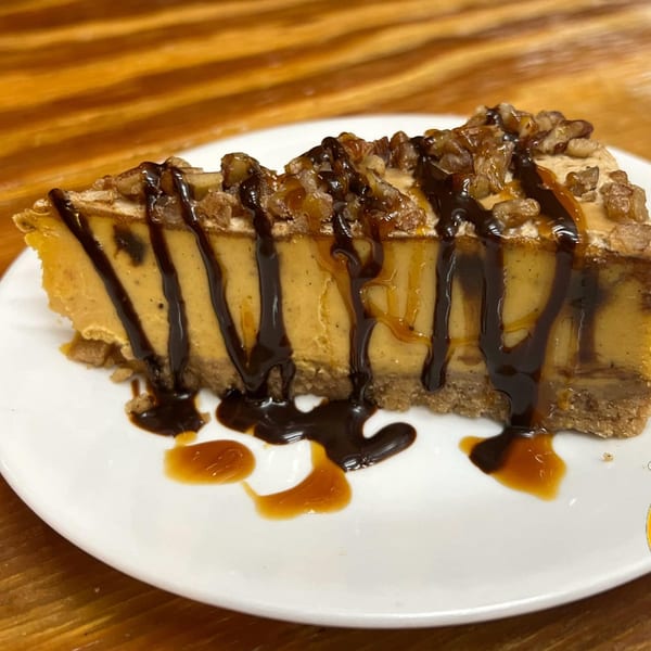 Slice of pumpkin cheesecake drizzled with chocolate and caramel, topped with walnuts on a white plate.