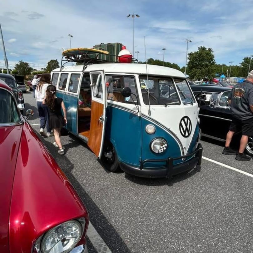 Blue and white Volkswagen van with open door, surfboard on roof, parked at an outdoor car show.